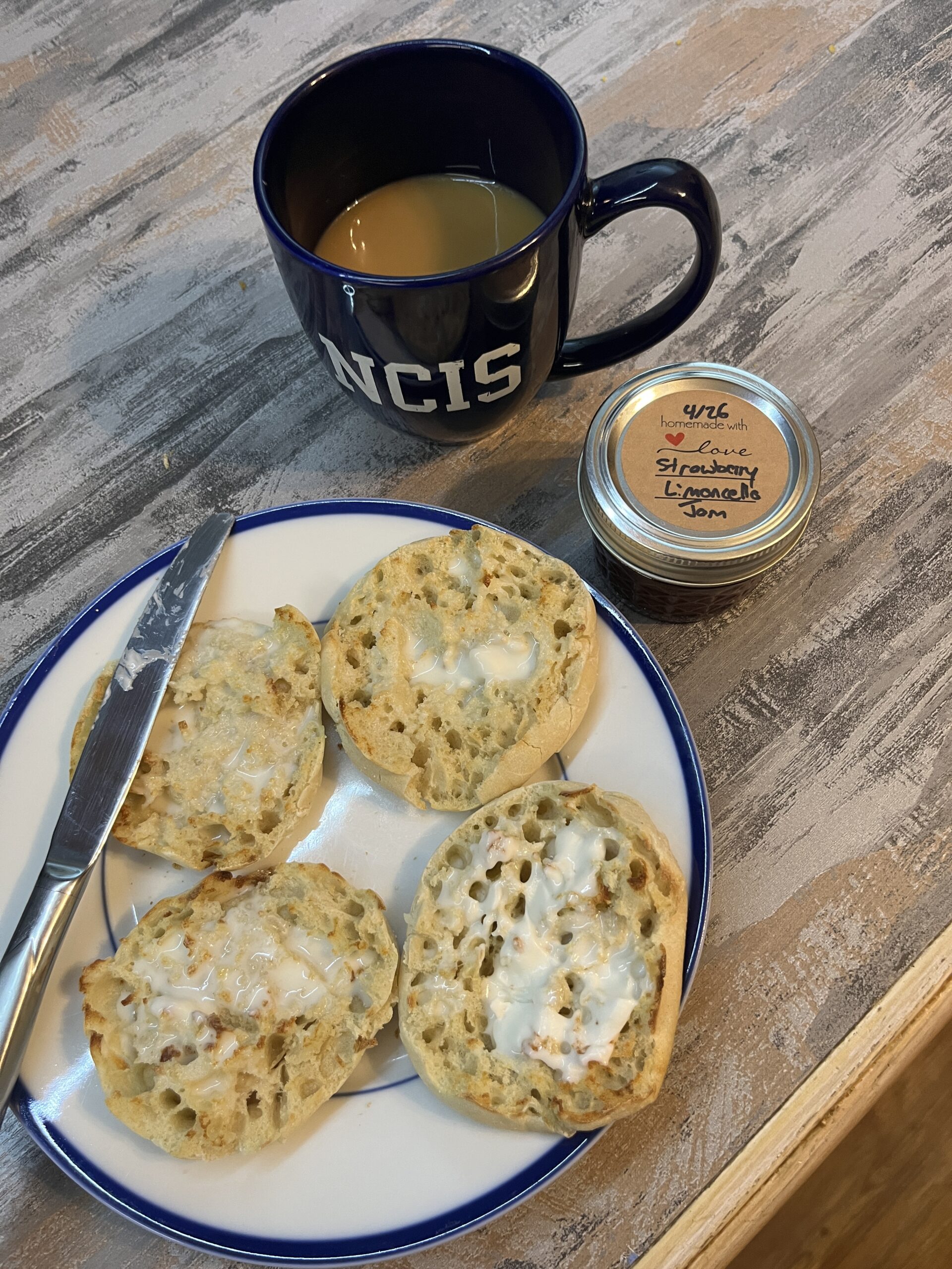 English muffins with butter on a plate with a jam jar and NCIS blue coffee mug